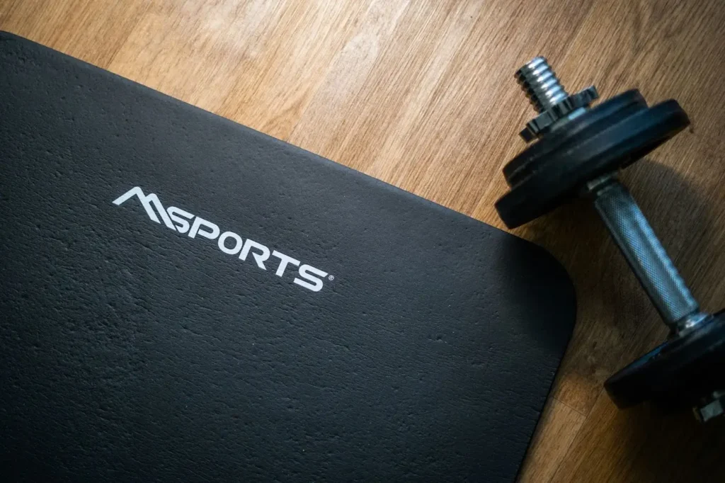 A dumbbell and a black workout mat on a wooden floor.