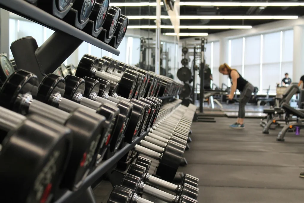 A close-up of a rack of weights at the gym. In the distance, a woman does a workout.
